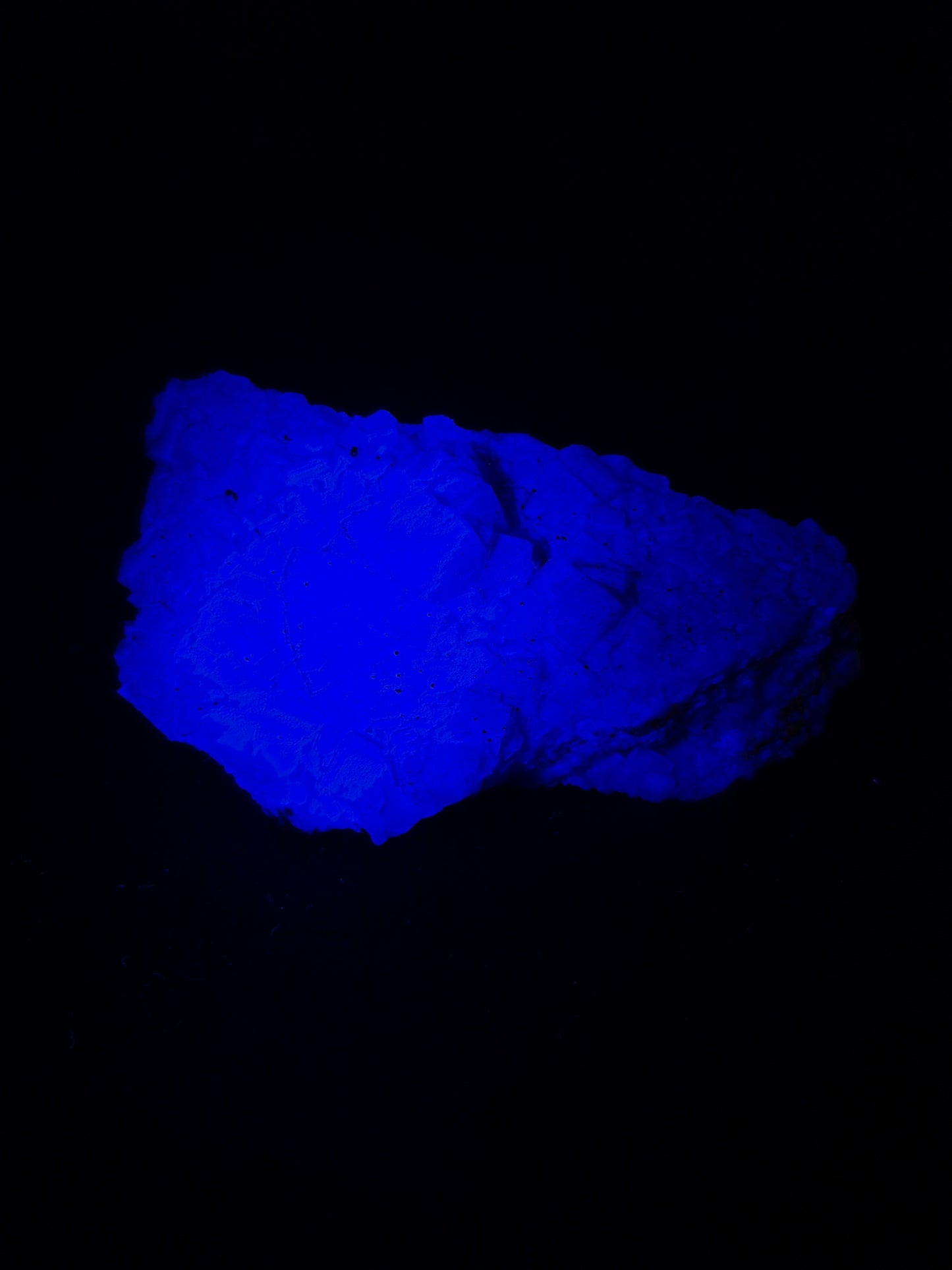 Raw black fluorite chunk photographed under ultraviolet light, glowing a vivid electric blue with visible natural fractures and crystalline texture, set against a dark background.