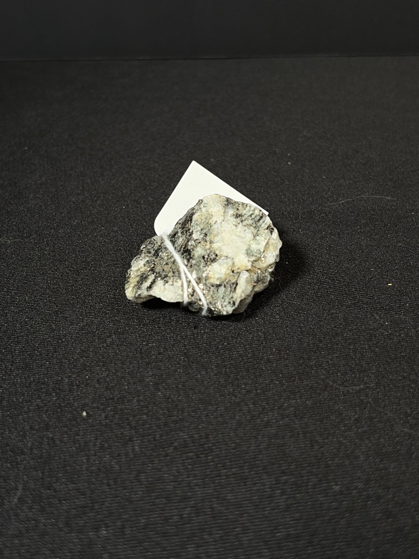 Raw rainbow moonstone mineral chunk resting on a black fabric surface, showing a rough natural texture with silvery-white and gray tones and subtle reflective shimmer. A small white tag is tied to the stone with string.