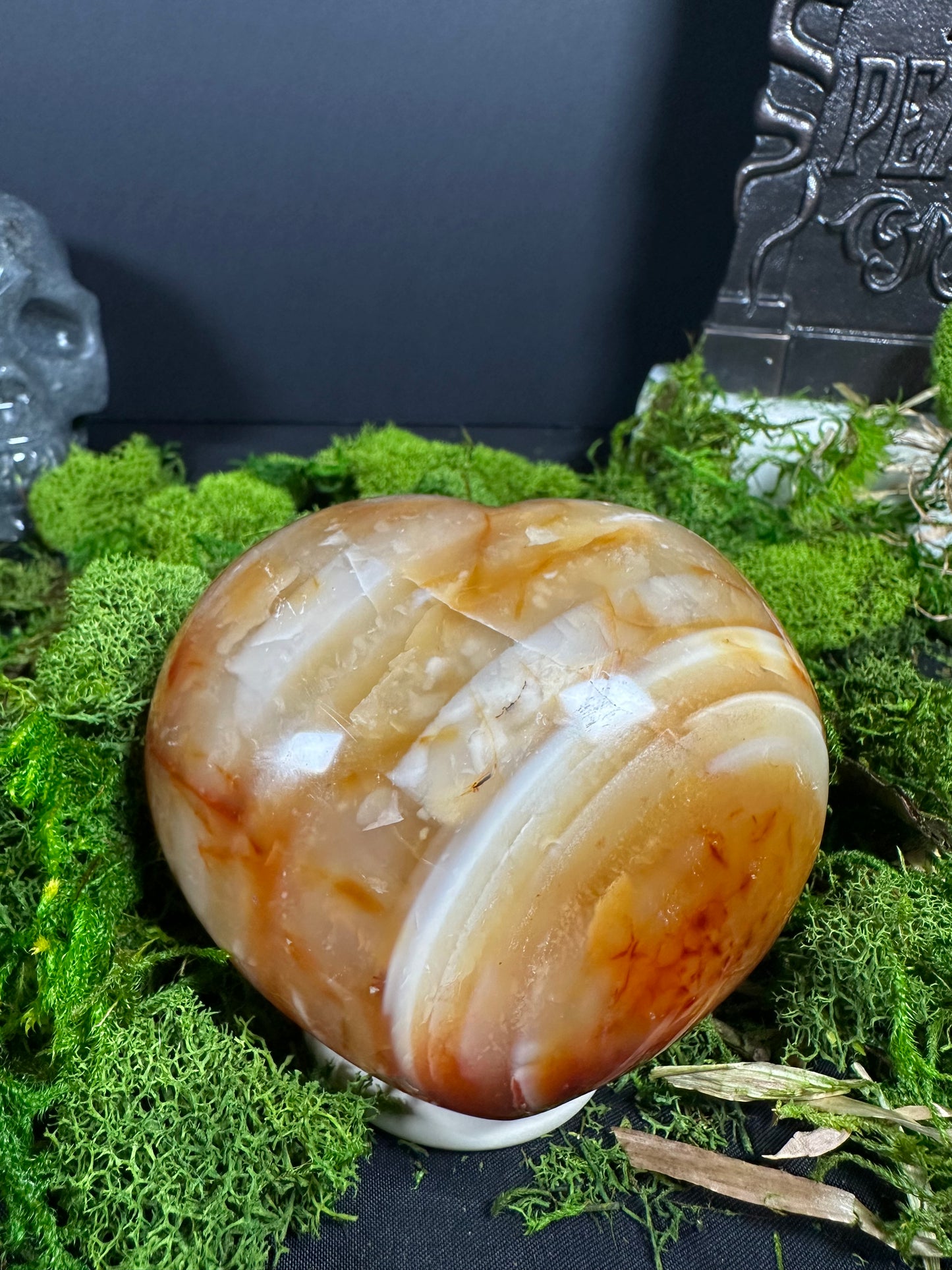 Large polished carnelian heart displayed upright on a white stand surrounded by vibrant green moss. The heart features creamy white, honey, and orange banding with translucent layers and a glossy finish, creating a warm, eye-catching display.