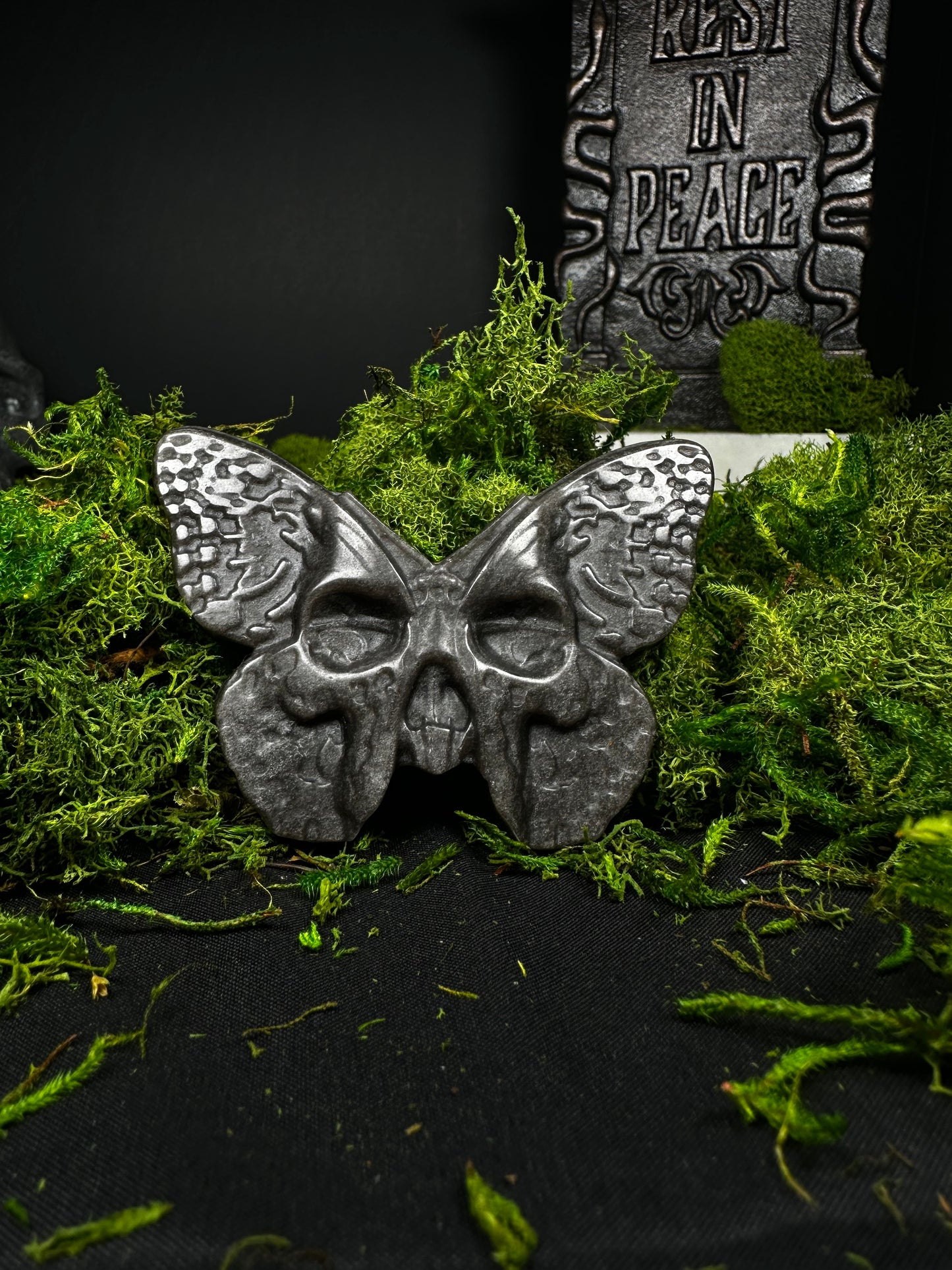 A polished obsidian carving shaped like a death’s-head moth rests on a bed of vibrant green moss against a black backdrop. The wings feature textured, engraved details, while the body forms a small skull with hollow eye sockets. A gray “Rest in Peace” tombstone stands softly blurred in the background, creating a gothic cemetery-inspired display that highlights the glossy black stone and intricate craftsmanship.
