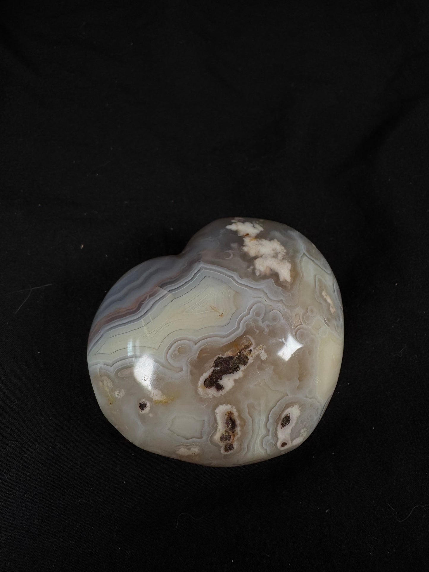 A single polished flower agate heart on a black fabric background. The heart features soft gray and cream banding with delicate white blossom formations, orbicular patterns, and a few natural druzy-lined cavities, creating a striking blend of floral and lace-like agate textures.