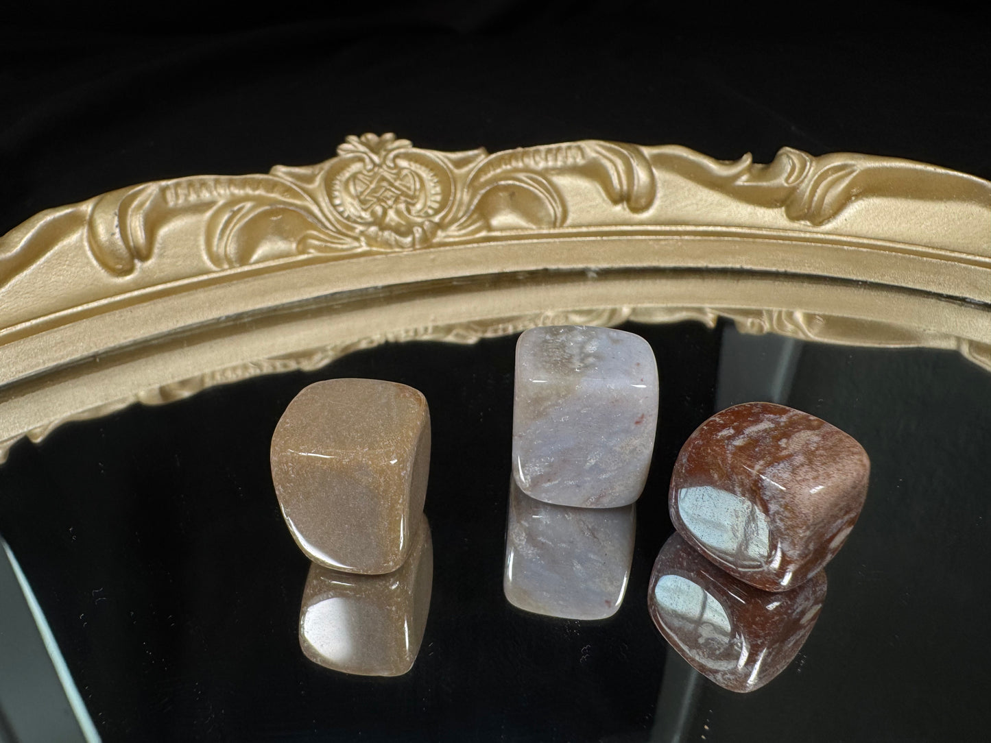 Polished Ocean Jasper cube tumble displayed on a black reflective surface with a decorative gold mirror tray. The cube features soft cream, beige, and muted blue-gray tones with natural orb-like patterns and subtle earthy inclusions. Its smooth, rounded edges and glossy finish highlight the unique colors and markings, while the reflective surface creates a mirrored view of the stone below.