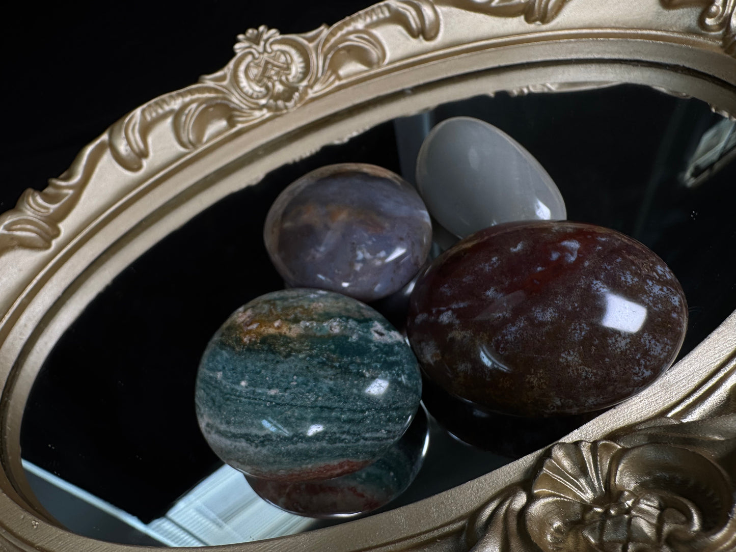 Collection of polished Ocean Jasper palm stones in assorted natural colors, including deep red, green, lavender, and light gray, displayed on an ornate vintage gold-framed mirror with glossy reflections against a black background.