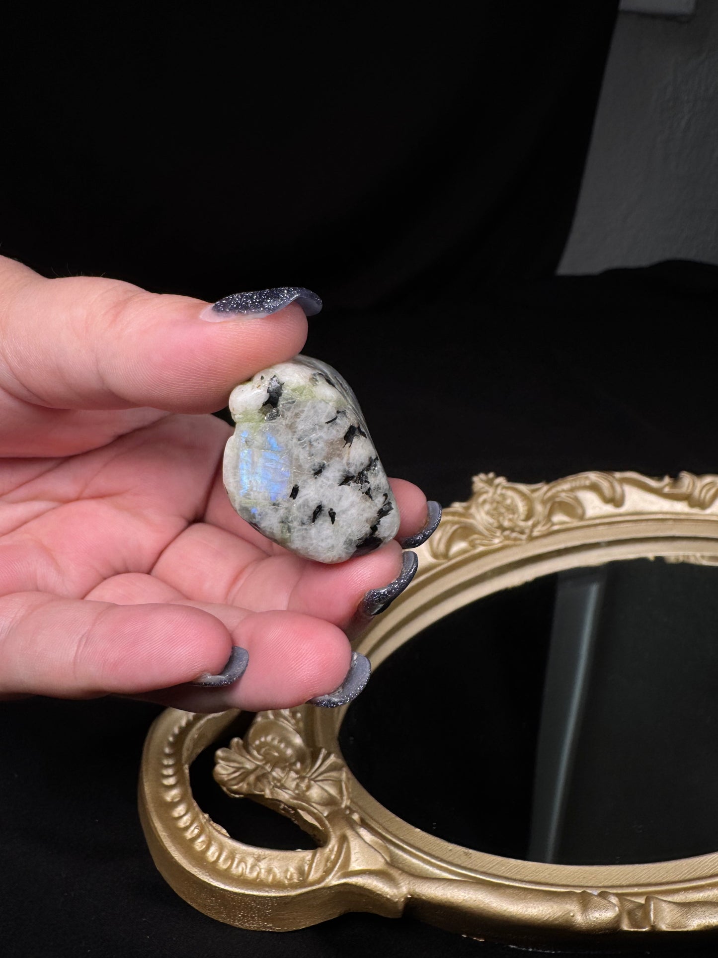Hand holding a polished rainbow moonstone tumble with black tourmaline inclusions. The stone displays a vivid blue flash on one side and is photographed against a black background above an ornate gold-framed mirror.