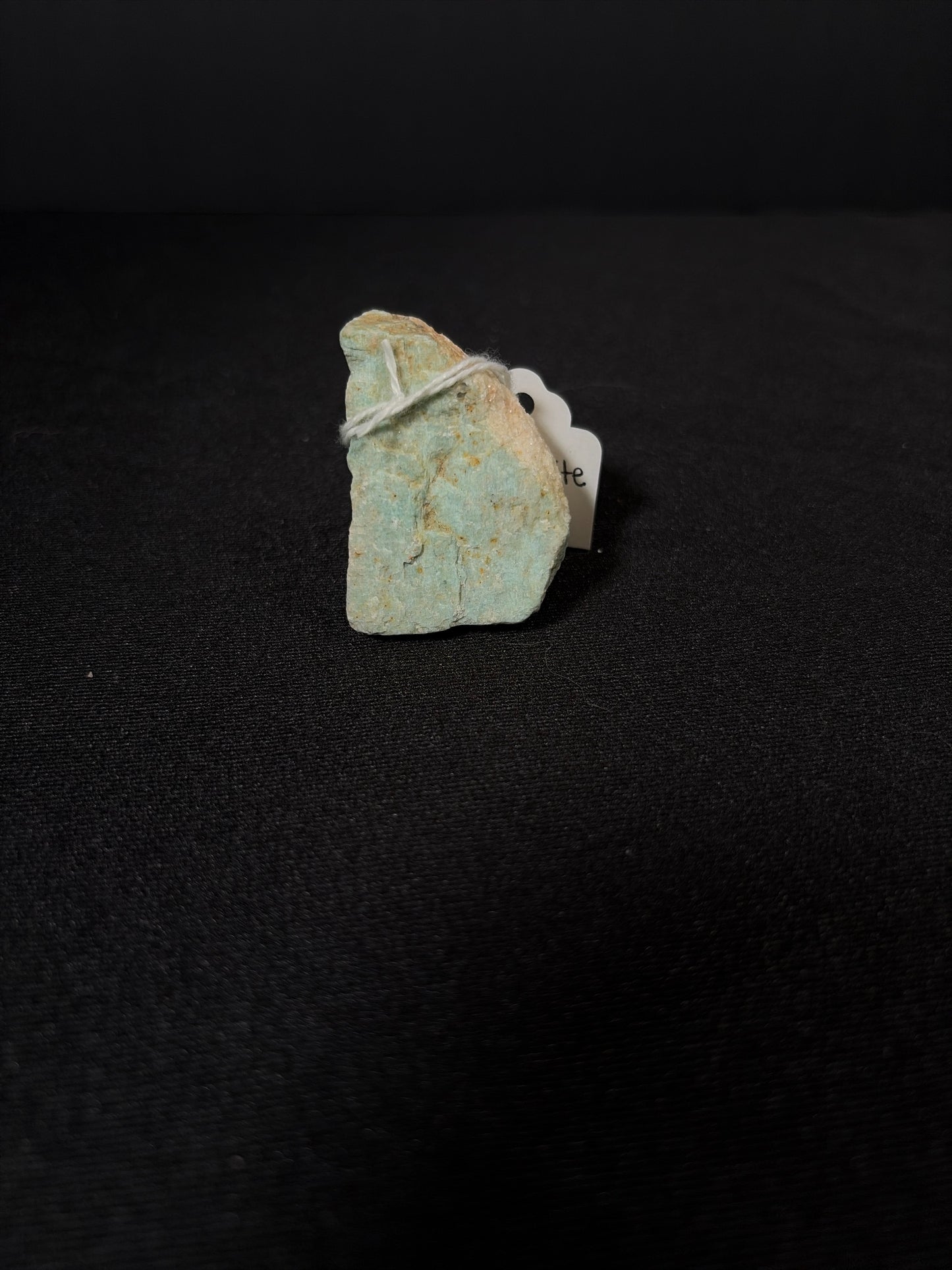 Raw Amazonite crystal chunk with a natural rough texture and pale turquoise-green color, lightly speckled with tan mineral inclusions. The stone is irregularly shaped and tied with a small piece of twine, resting upright against a black fabric background with a small tag partially visible behind it.