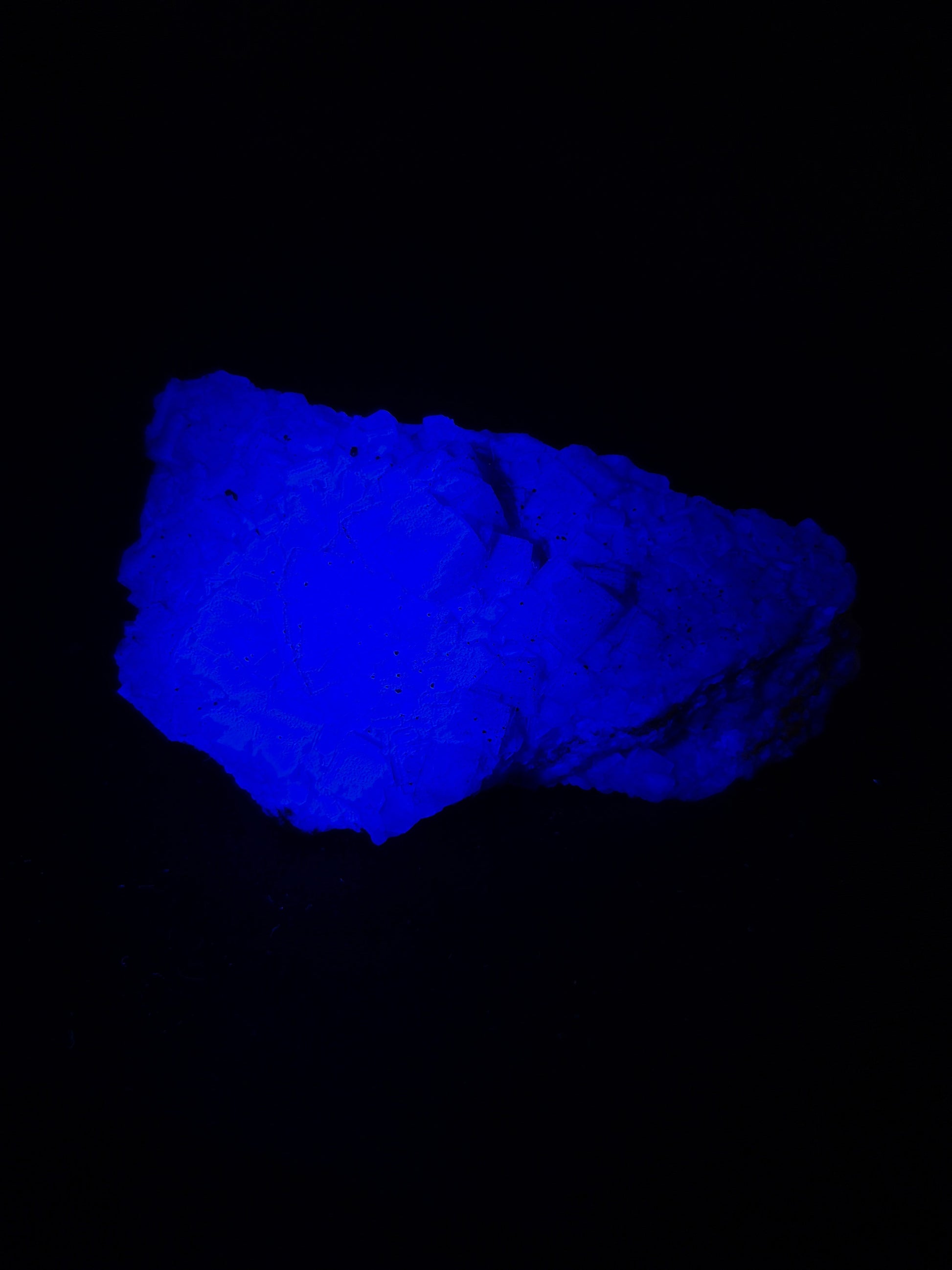 Raw black fluorite chunk photographed under ultraviolet light, glowing a vivid electric blue with visible natural fractures and crystalline texture, set against a dark background.