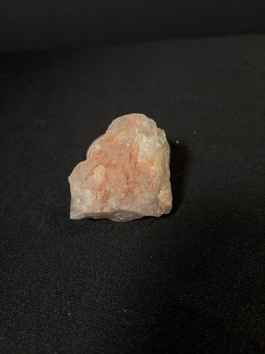 Raw strawberry quartz crystal chunk resting on a black fabric surface, featuring a rough natural shape with translucent milky quartz and soft reddish-pink mineral inclusions throughout.