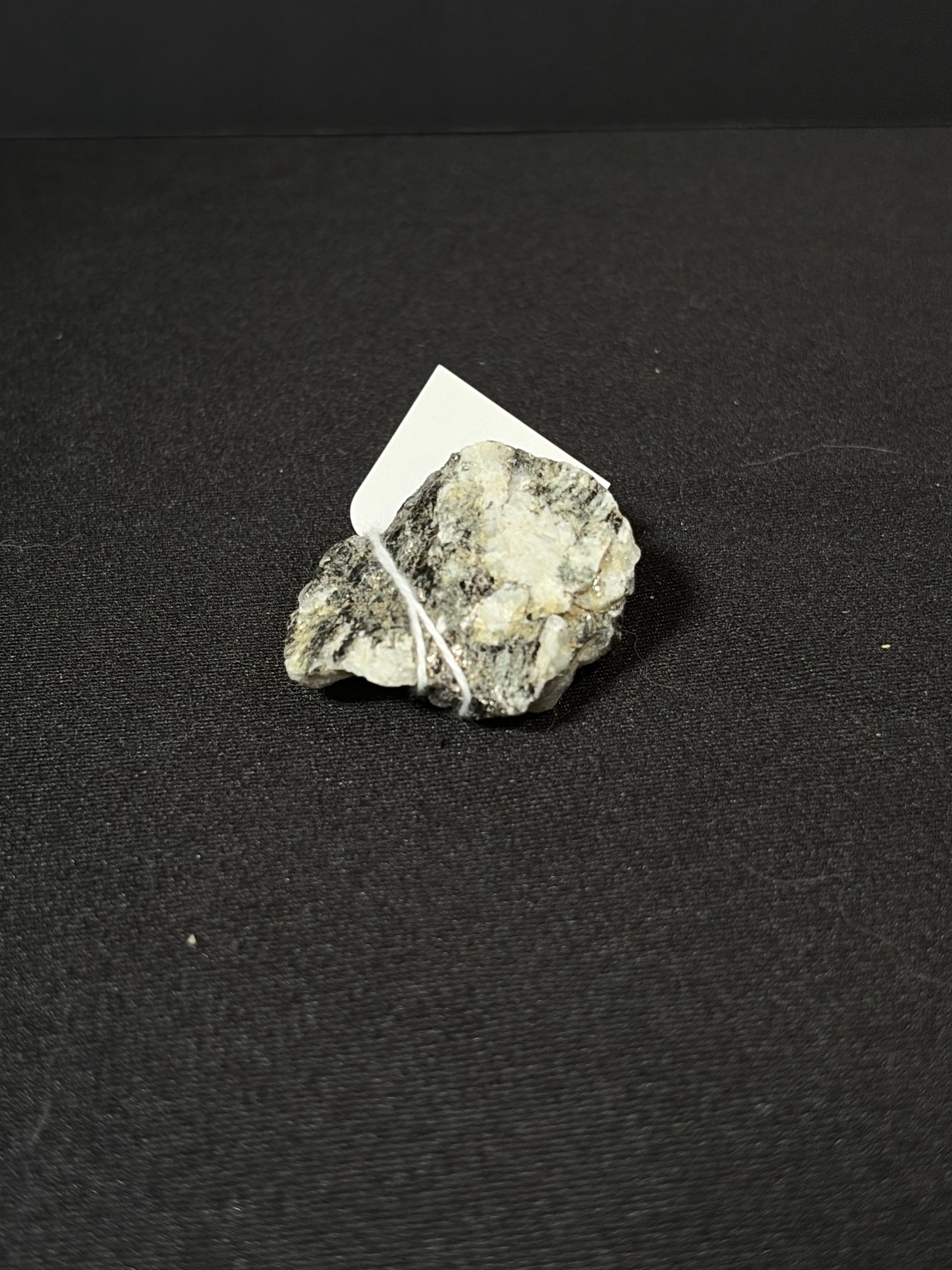 Raw rainbow moonstone mineral chunk resting on a black fabric surface, showing a rough natural texture with silvery-white and gray tones and subtle reflective shimmer. A small white tag is tied to the stone with string.