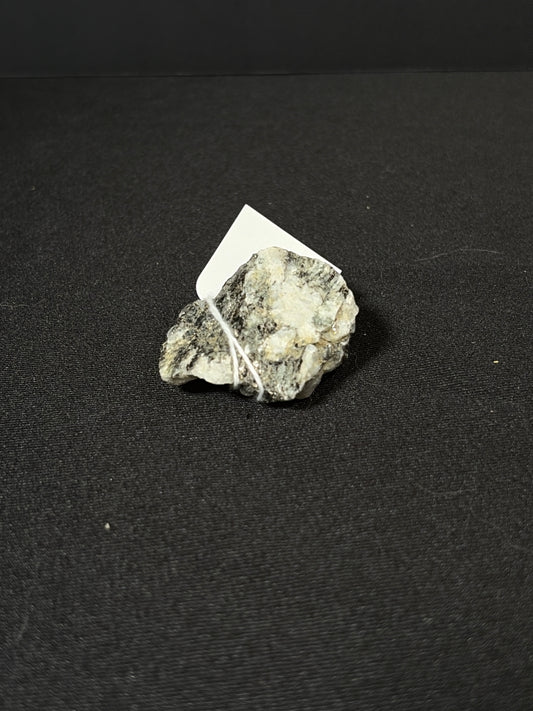 Raw rainbow moonstone mineral chunk resting on a black fabric surface, showing a rough natural texture with silvery-white and gray tones and subtle reflective shimmer. A small white tag is tied to the stone with string.