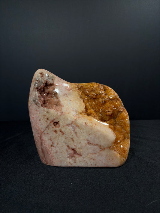 Polished pink amethyst and citrine freeform crystal standing upright on a black fabric surface against a dark background. The stone features soft blush and cream tones with natural veining, a sparkling pink amethyst druzy pocket near the top left, and a golden honey-colored citrine crystal cluster along the right side.