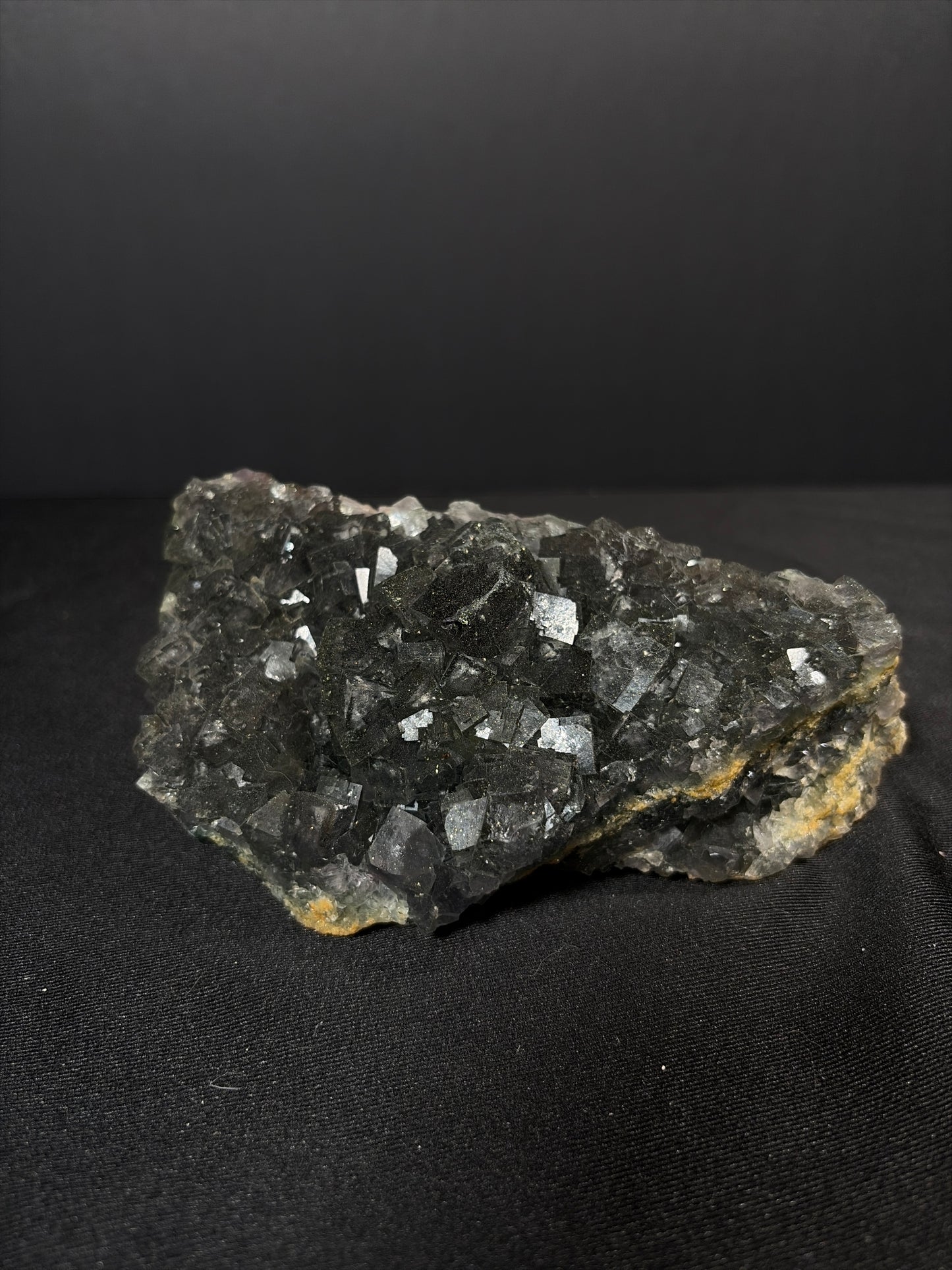 Raw black fluorite chunk resting on a black fabric surface, featuring dark smoky-black cubic crystals with natural texture and subtle reflective faces, photographed against a dark gradient background.