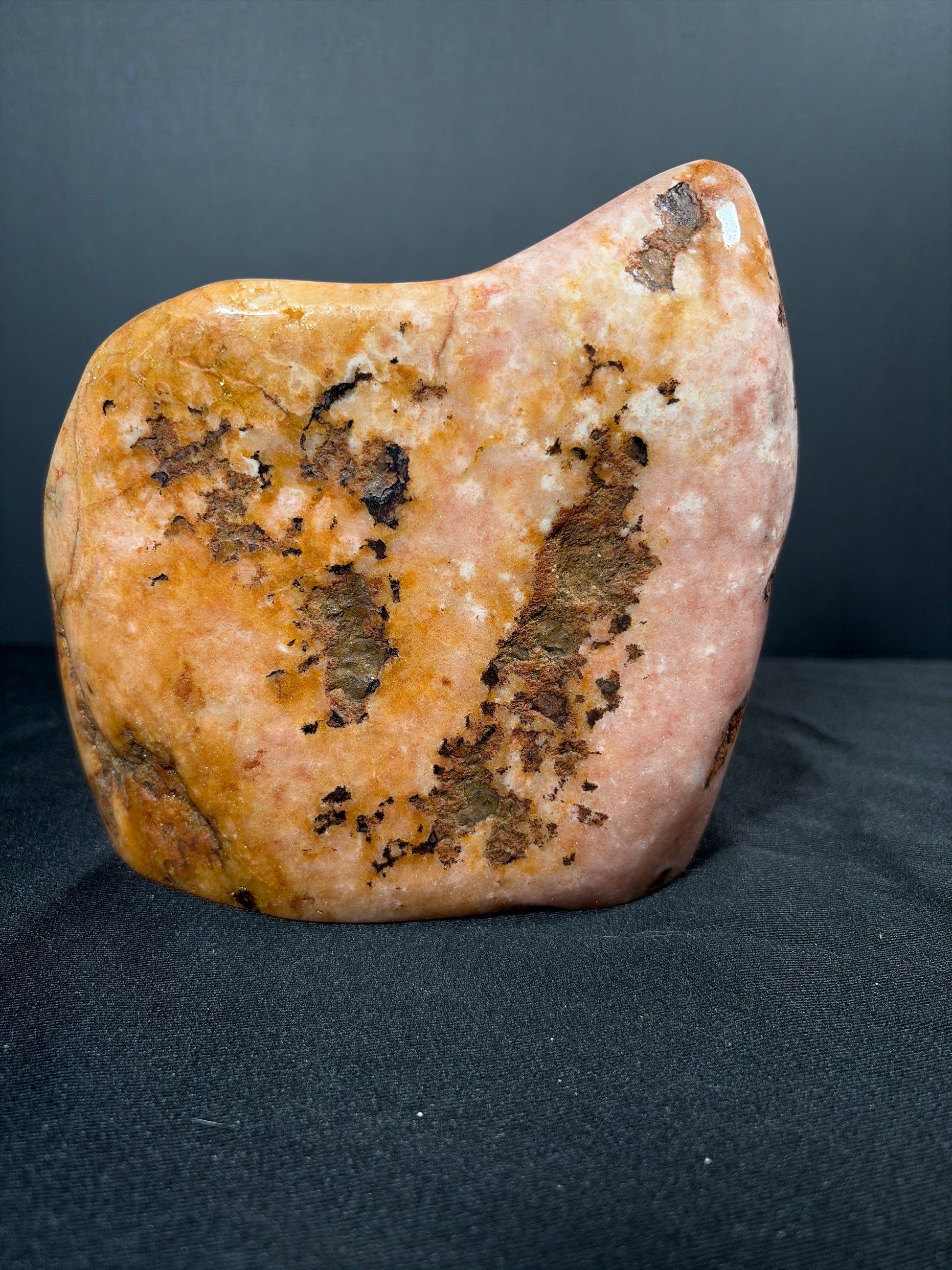 Polished pink amethyst and citrine freeform crystal displayed upright on a black fabric surface against a dark background. The stone features soft blush-pink and warm golden tones with natural marbling, along with scattered darker mineral inclusions that create organic patterns across the surface.