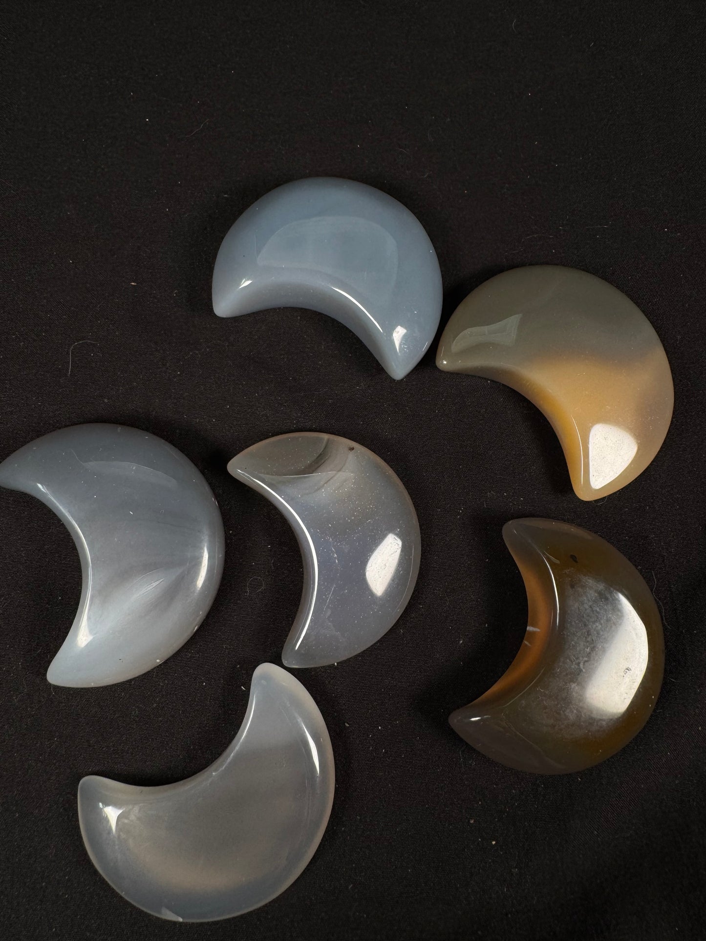 Six polished agate crescent moon carvings displayed on a black fabric background. The moons range in color from soft gray-blue and translucent gray to warm honey-brown and deep smoky brown, with smooth glossy finishes that reflect the light. Each crescent showcases the natural color variations and subtle banding unique to agate.