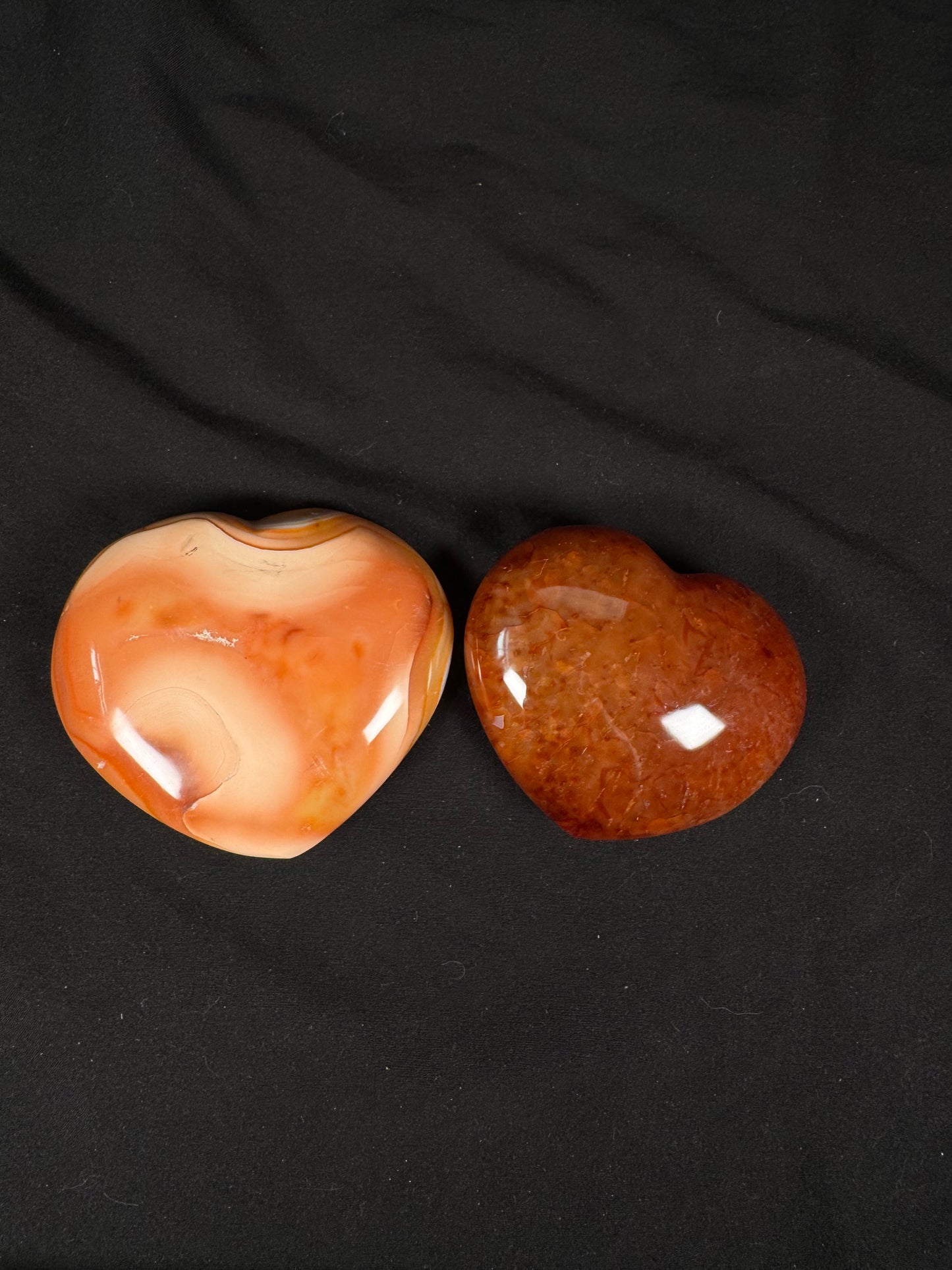 Two polished carnelian crystal hearts displayed on a black fabric background. One heart features soft peach, cream, and orange banding with translucent areas, while the other has a rich reddish-orange color with natural internal patterns and a glossy finish.