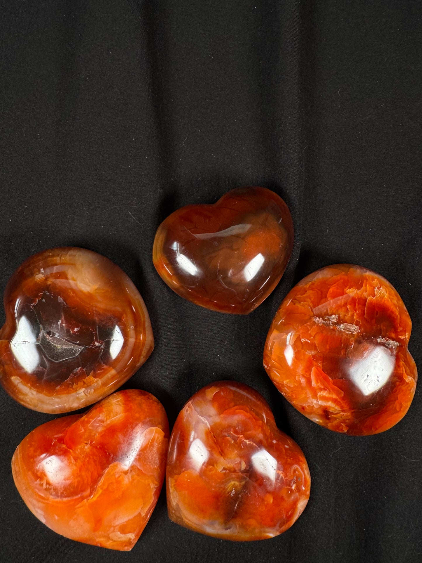 Five polished carnelian crystal hearts arranged on a black fabric background. Each heart displays unique shades of fiery orange, deep red, amber, and translucent white banding, with natural inclusions and a high-polish shine.