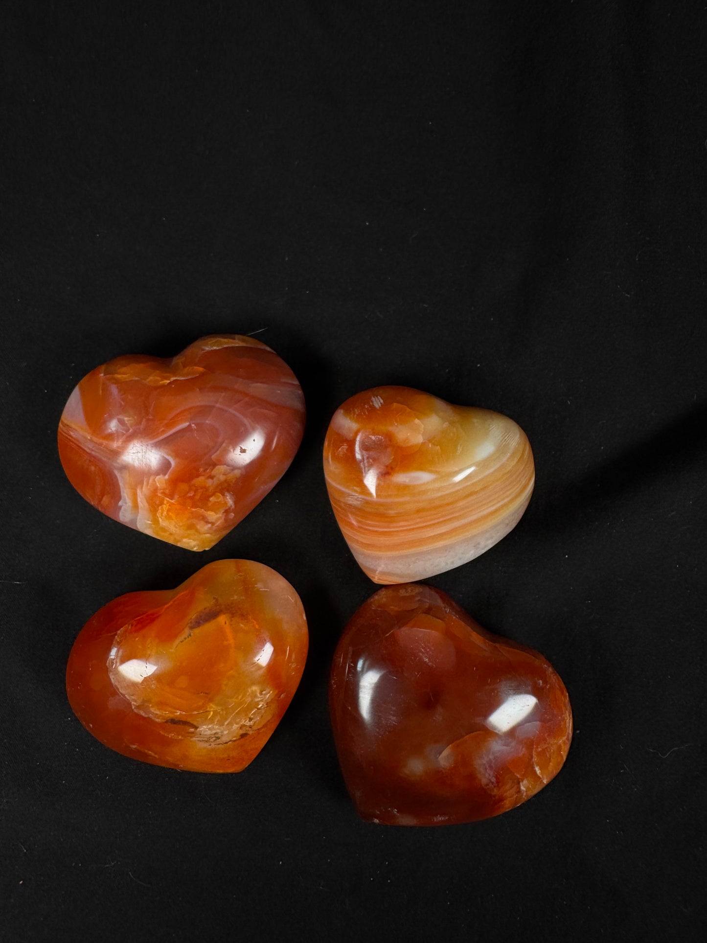 Four polished carnelian crystal hearts arranged on a black fabric background. The hearts showcase warm orange, red, amber, and cream tones with striking natural banding, swirls, and translucent sections, each with a smooth glossy finish.