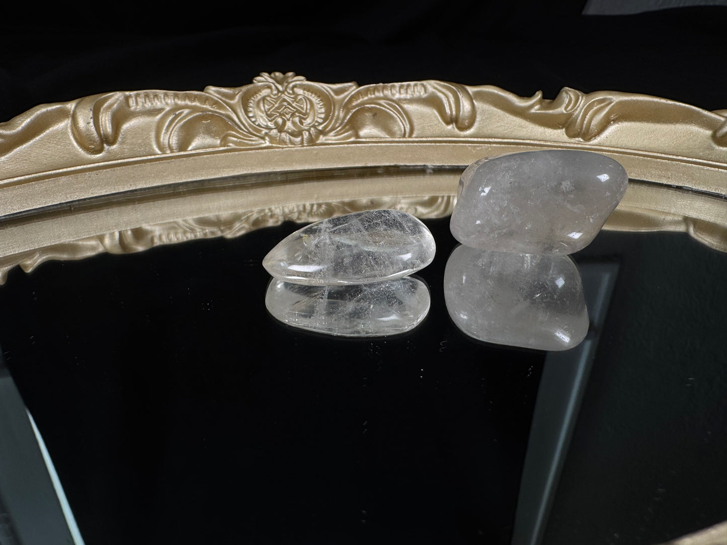 Two polished clear quartz tumbles displayed on a glossy black reflective surface with an ornate antique-style gold mirror frame in the background. One crystal is nearly transparent with visible wispy inclusions, while the other has a soft cloudy translucence. Their smooth, rounded surfaces and mirrored reflections highlight the natural beauty and unique character of each stone.