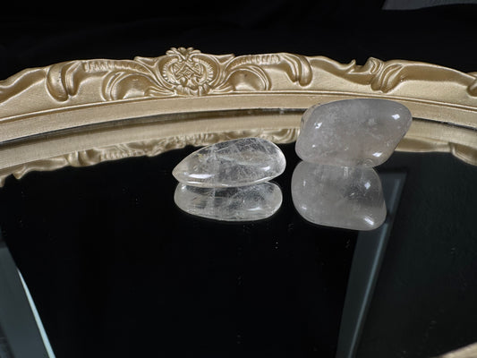 Two polished clear quartz tumbles displayed on a glossy black reflective surface with an ornate antique-style gold mirror frame in the background. One crystal is nearly transparent with visible wispy inclusions, while the other has a soft cloudy translucence. Their smooth, rounded surfaces and mirrored reflections highlight the natural beauty and unique character of each stone.