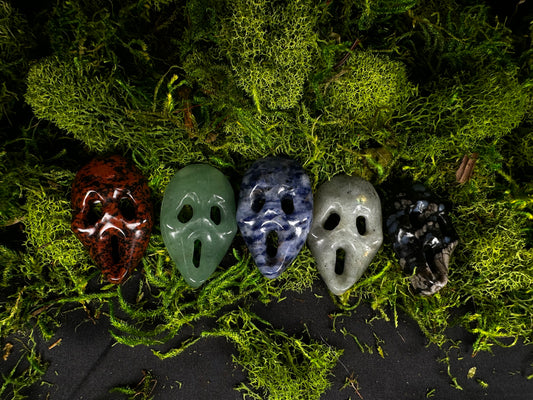Five miniature crystal ghostface-style mask carvings displayed on a bed of vibrant green moss. The masks feature a variety of natural stones, including deep mahogany obsidian, green aventurine, blue sodalite, gray labradorite, and black snowflake obsidian, each showcasing unique colors and natural patterns. The spooky miniature carvings are arranged in a row against a dark background, highlighting their polished finish and intricate facial details.