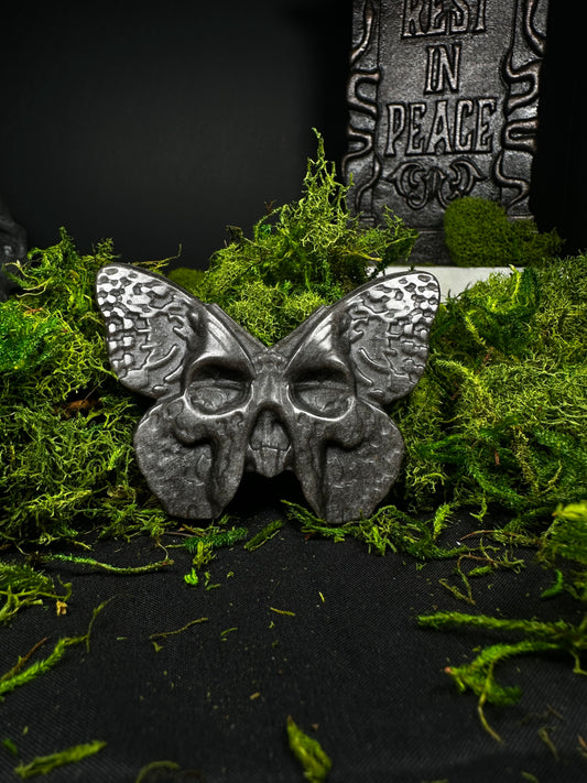A polished obsidian carving shaped like a death’s-head moth rests on a bed of vibrant green moss against a black backdrop. The wings feature textured, engraved details, while the body forms a small skull with hollow eye sockets. A gray “Rest in Peace” tombstone stands softly blurred in the background, creating a gothic cemetery-inspired display that highlights the glossy black stone and intricate craftsmanship.