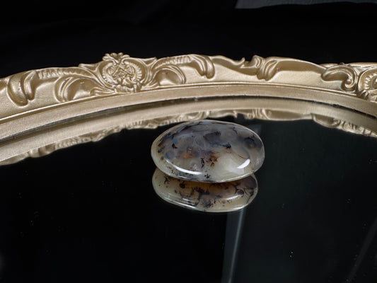 Polished dendritic agate tumble with a translucent milky white base featuring striking black, branch-like dendritic inclusions and soft gray clouding throughout. The smooth oval stone is displayed on a reflective black surface, creating a mirror-like reflection, with an ornate antique gold frame in the background that highlights the agate’s natural patterns.