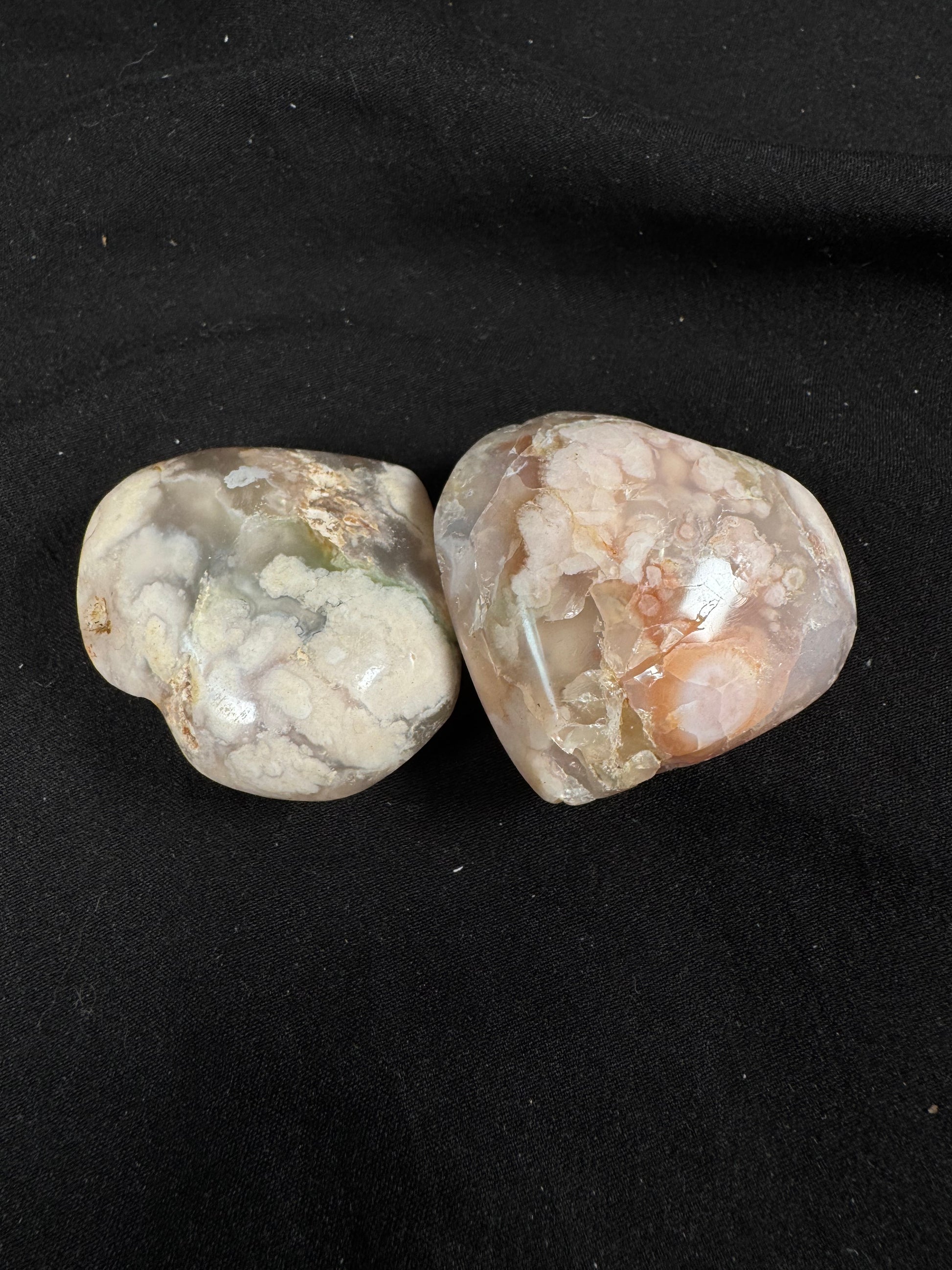 Two polished flower agate hearts resting on a black fabric background. Both hearts display translucent cream, peach, and soft gray tones with intricate white floral inclusions. One heart features natural internal fractures and unique crystalline patterns that make it one of a kind.