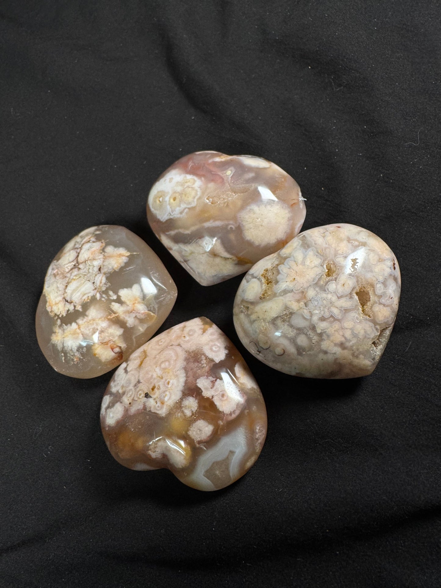 Four polished flower agate hearts arranged on a black fabric background. Each heart showcases unique combinations of translucent gray, beige, blush pink, and cream with signature blossom-like plumes and orbicular patterns, highlighting the natural beauty and individuality of every piece.
