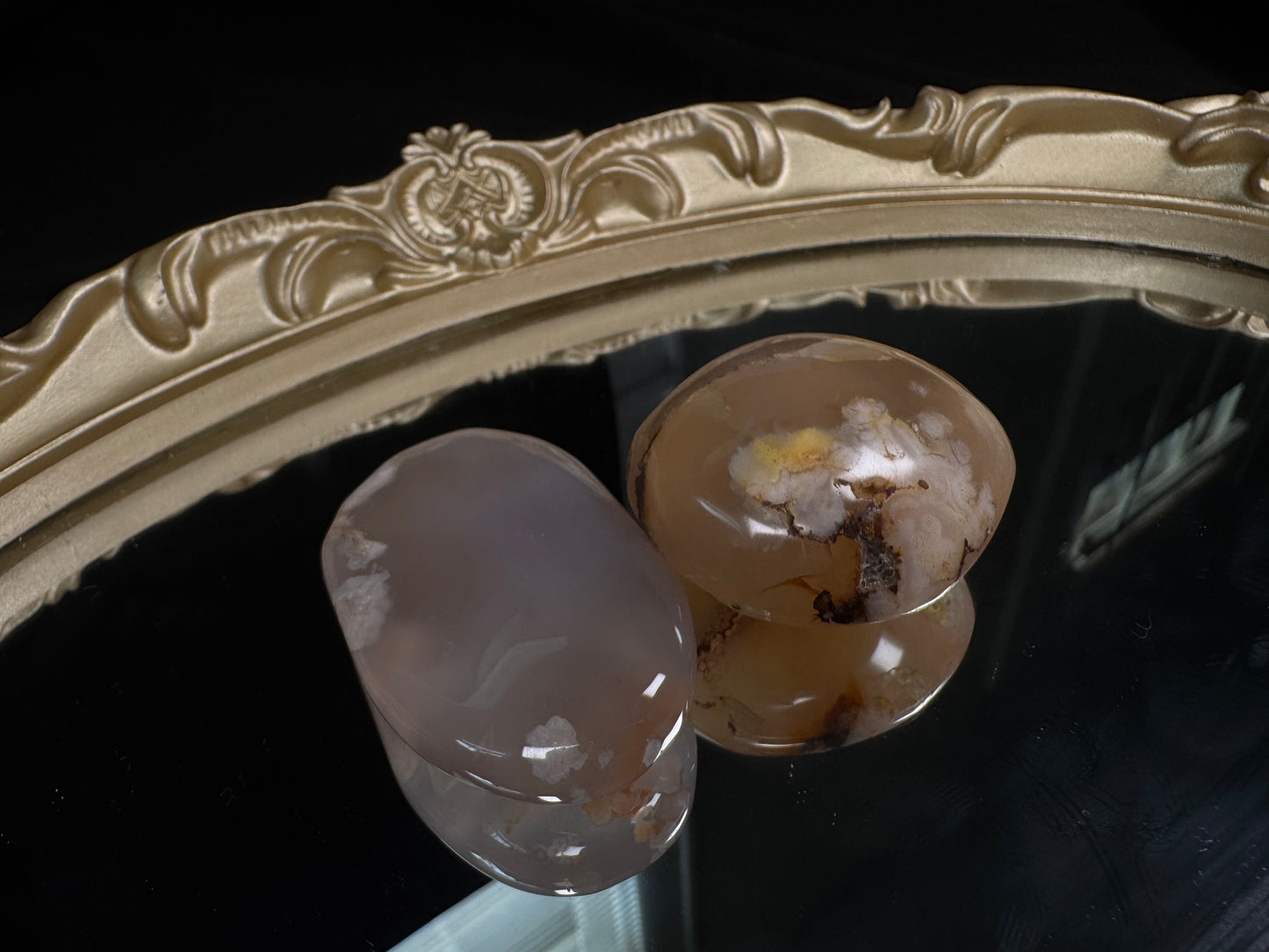 Two polished flower agate palm stones displayed on a vintage gold-framed mirror against a black background. The left stone is soft translucent gray with delicate white blossom-like plumes, while the right stone features warm peach and honey tones with prominent white flower formations, golden accents, and natural brown dendritic inclusions. Both palm stones have a smooth, glossy finish that highlights their unique floral patterns.