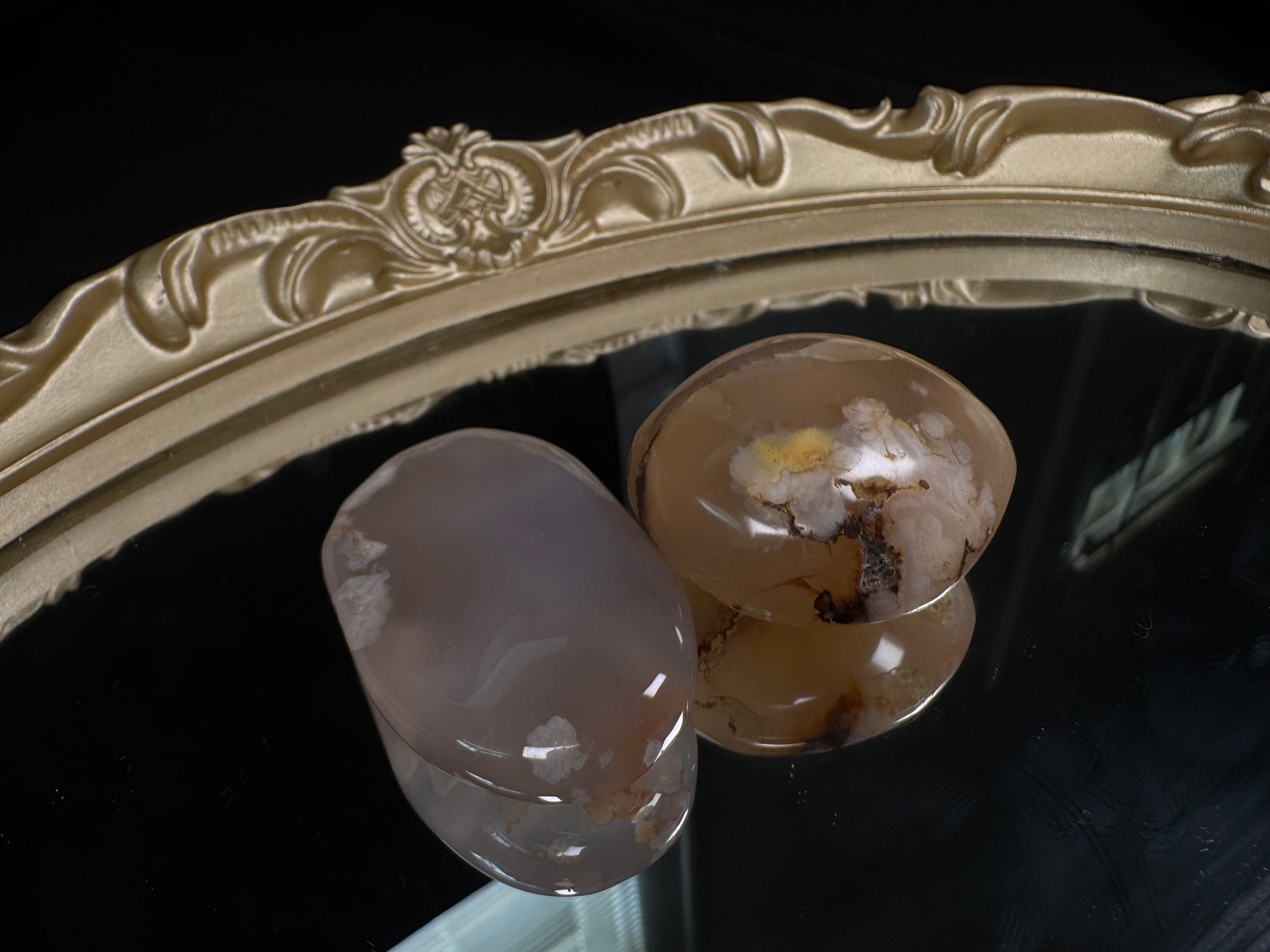 Two polished flower agate palm stones displayed on a vintage gold-framed mirror against a black background. The left stone is soft translucent gray with delicate white blossom-like plumes, while the right stone features warm peach and honey tones with prominent white flower formations, golden accents, and natural brown dendritic inclusions. Both palm stones have a smooth, glossy finish that highlights their unique floral patterns.