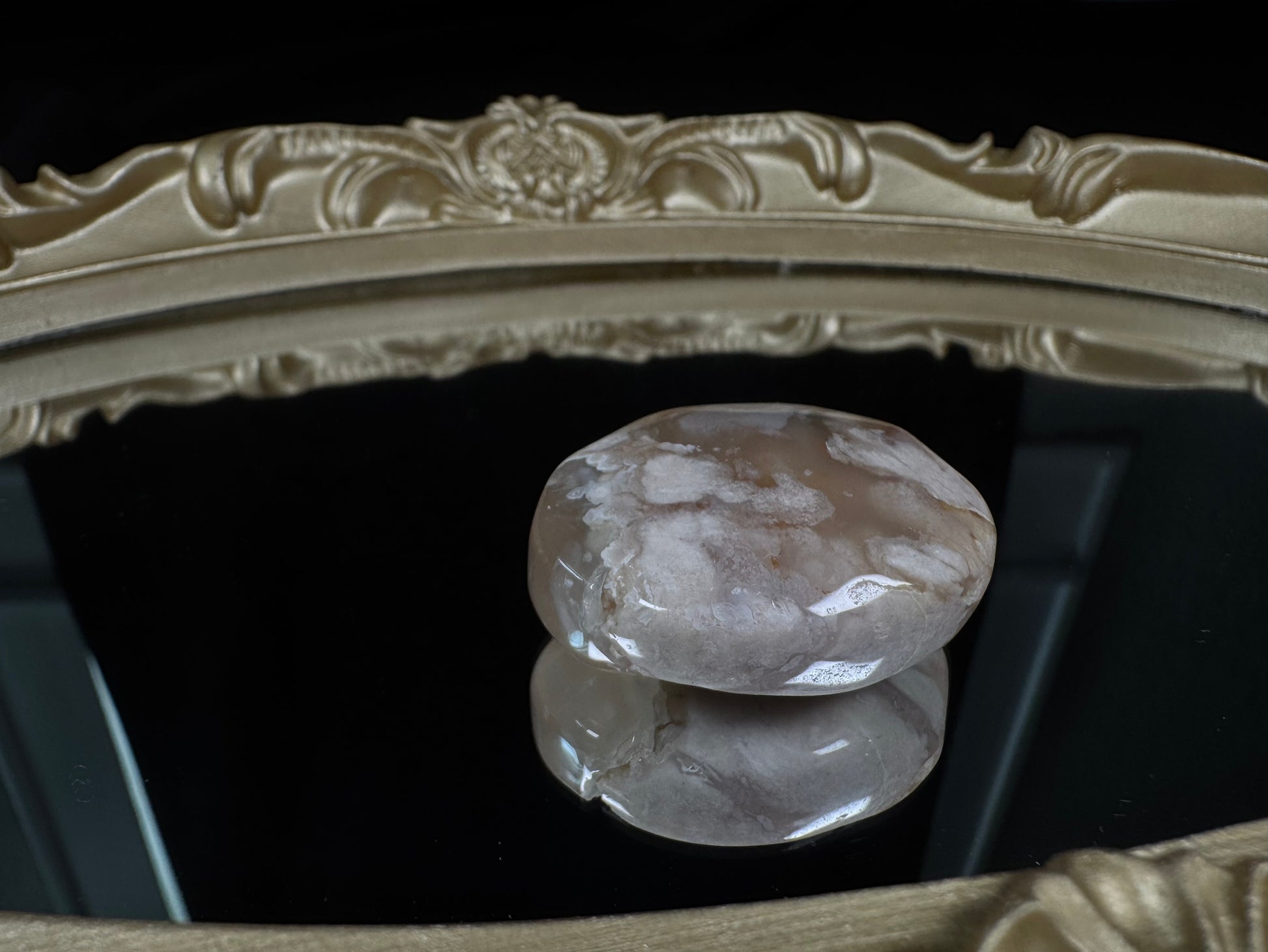 Polished flower agate tumble with soft blush pink and translucent cream tones, featuring white, cloud-like floral plume patterns throughout. Displayed on a glossy black reflective surface within an ornate antique-style gold frame, highlighting the stone’s smooth, rounded shape and natural botanical-like inclusions.