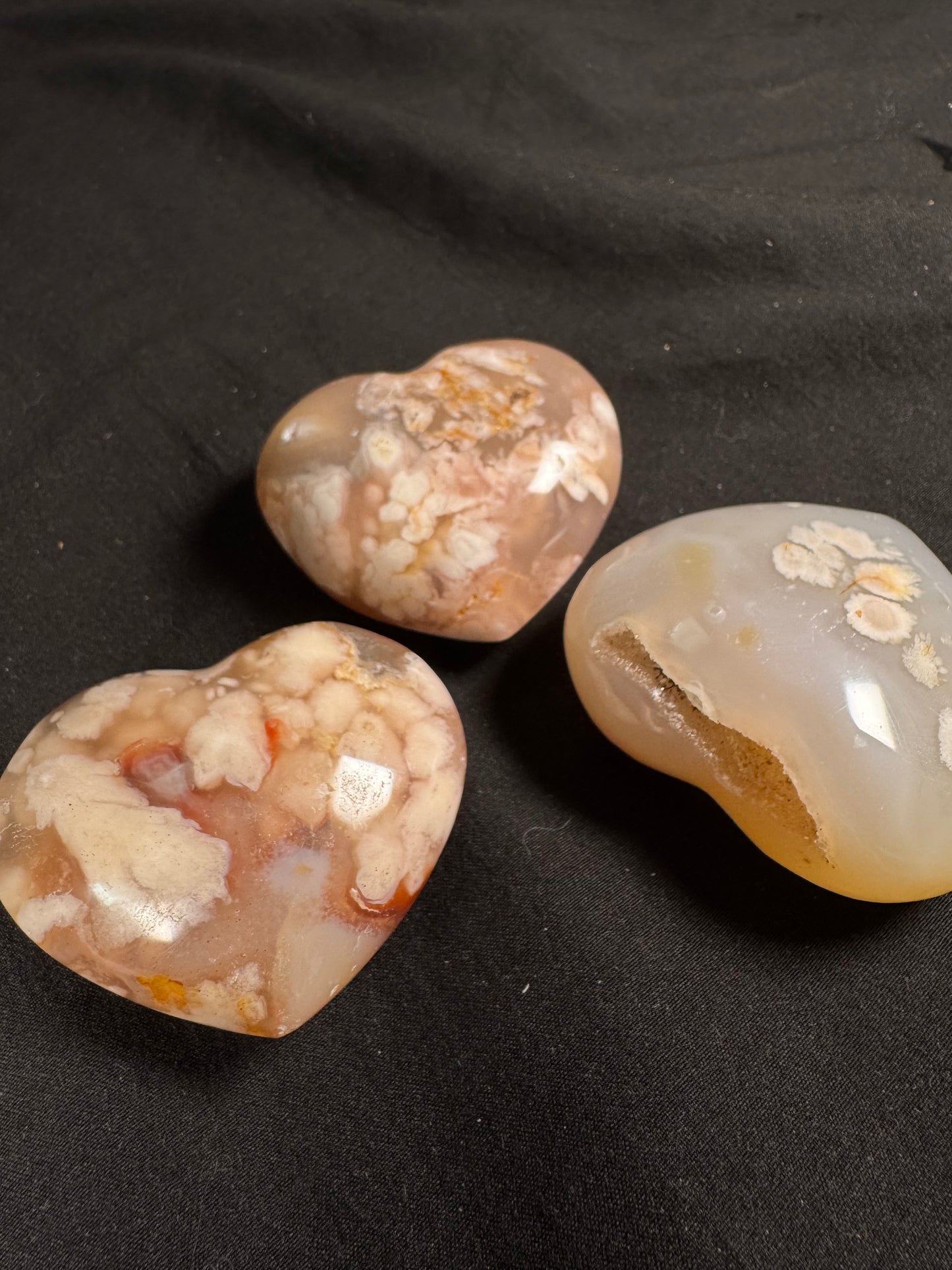 Three polished flower agate hearts displayed on a black fabric background. The hearts feature soft blush pink, cream, and translucent chalcedony with distinctive white, flower-like plume formations. One heart includes a natural druzy pocket along the edge, adding sparkling crystal texture.