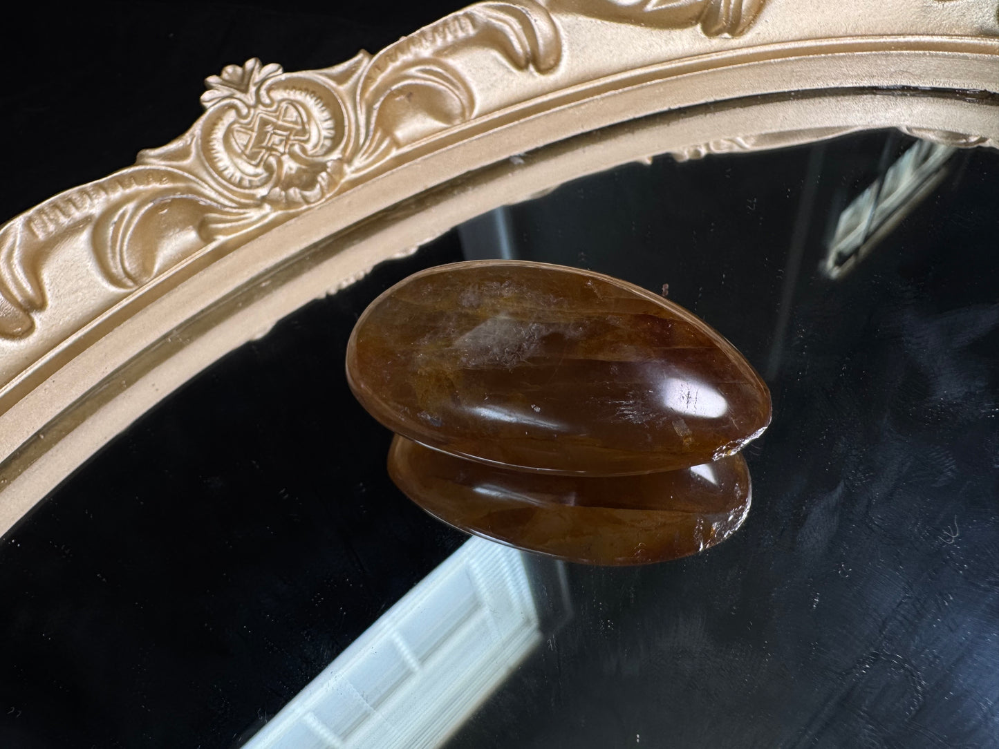 Polished golden healer quartz palm stone with warm golden-brown and honey-colored translucent tones, displaying natural internal veils and subtle mineral inclusions. The smooth oval stone rests on a black reflective surface beneath an ornate vintage-style gold mirror frame, highlighting its glossy finish and gentle color variations.