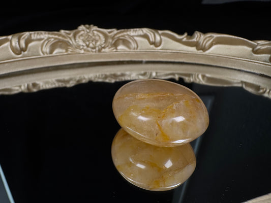 Polished Golden Healer palm stone with warm golden-yellow mineral inclusions suspended in translucent quartz. The smooth, oval-shaped stone is photographed on a black mirrored surface that creates a clear reflection, with an ornate antique-style gold frame softly blurred in the background, highlighting the stone’s natural clarity and glowing golden patterns.