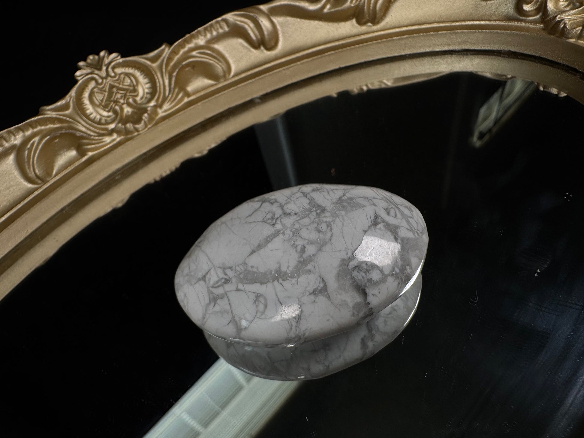 Polished howlite palm stone with a smooth oval shape, featuring a white to light gray base crossed by delicate gray veining in a natural marbled pattern. The stone is displayed on a black reflective surface beneath an ornate antique-style gold mirror frame, highlighting its glossy finish and unique mineral patterns.