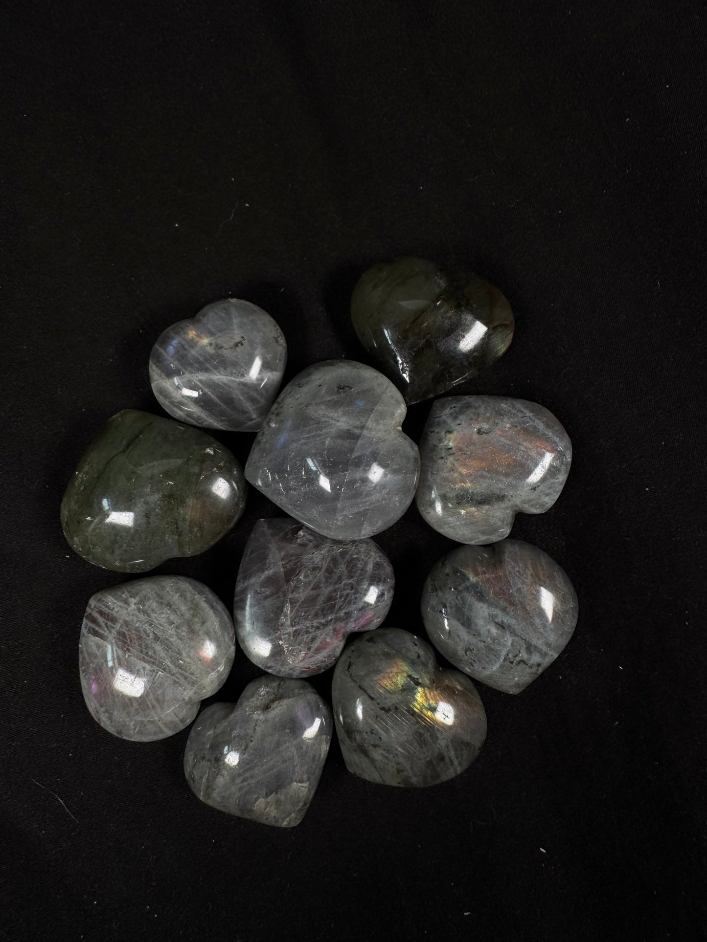 A collection of polished labradorite heart carvings arranged on a black fabric background. The hearts range from light gray to deep charcoal with natural black veining and inclusions, several displaying flashes of blue, gold, green, orange, and purple that shimmer in the light.