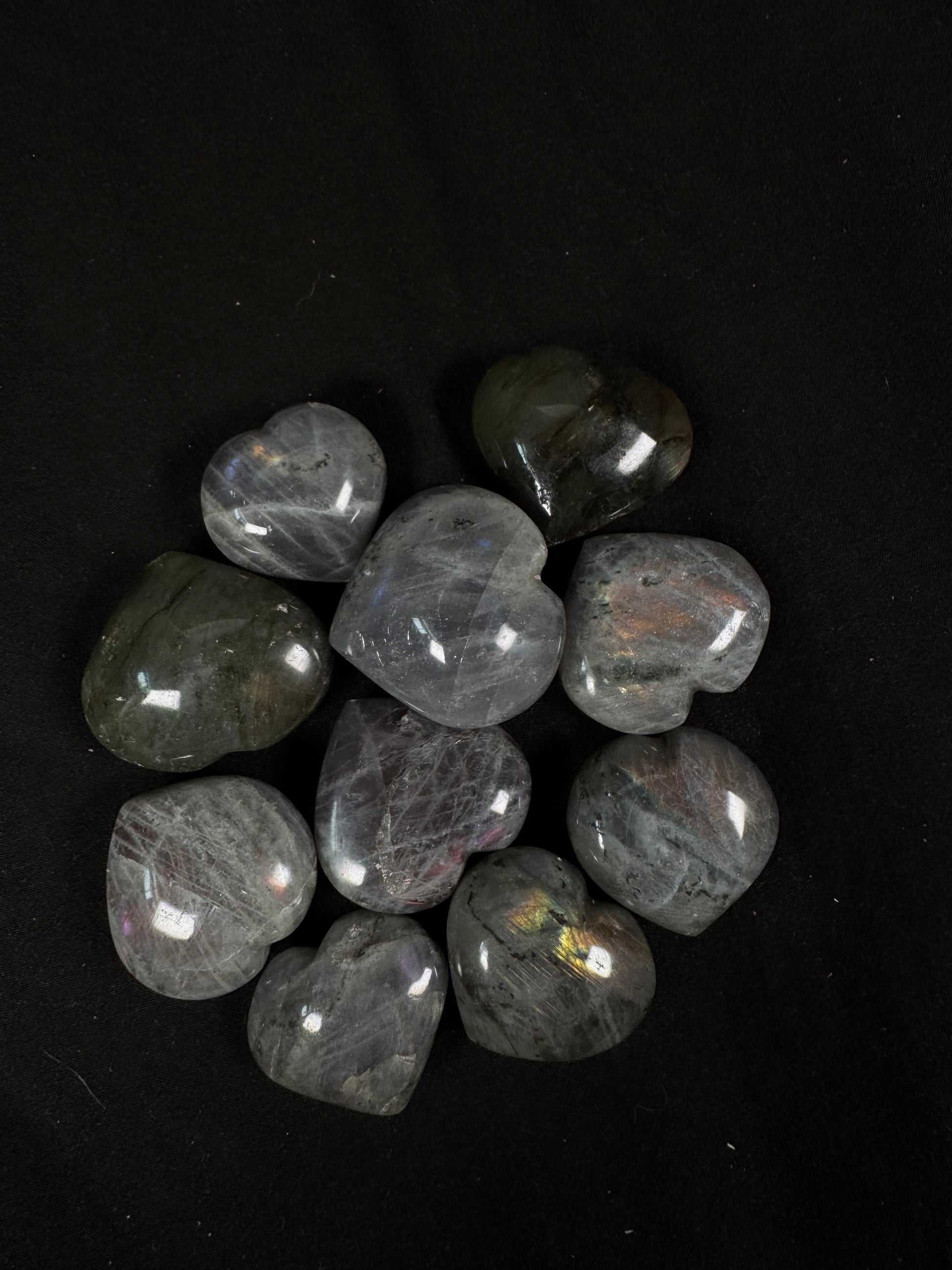 A collection of polished labradorite heart carvings arranged on a black fabric background. The hearts range from light gray to deep charcoal with natural black veining and inclusions, several displaying flashes of blue, gold, green, orange, and purple that shimmer in the light.