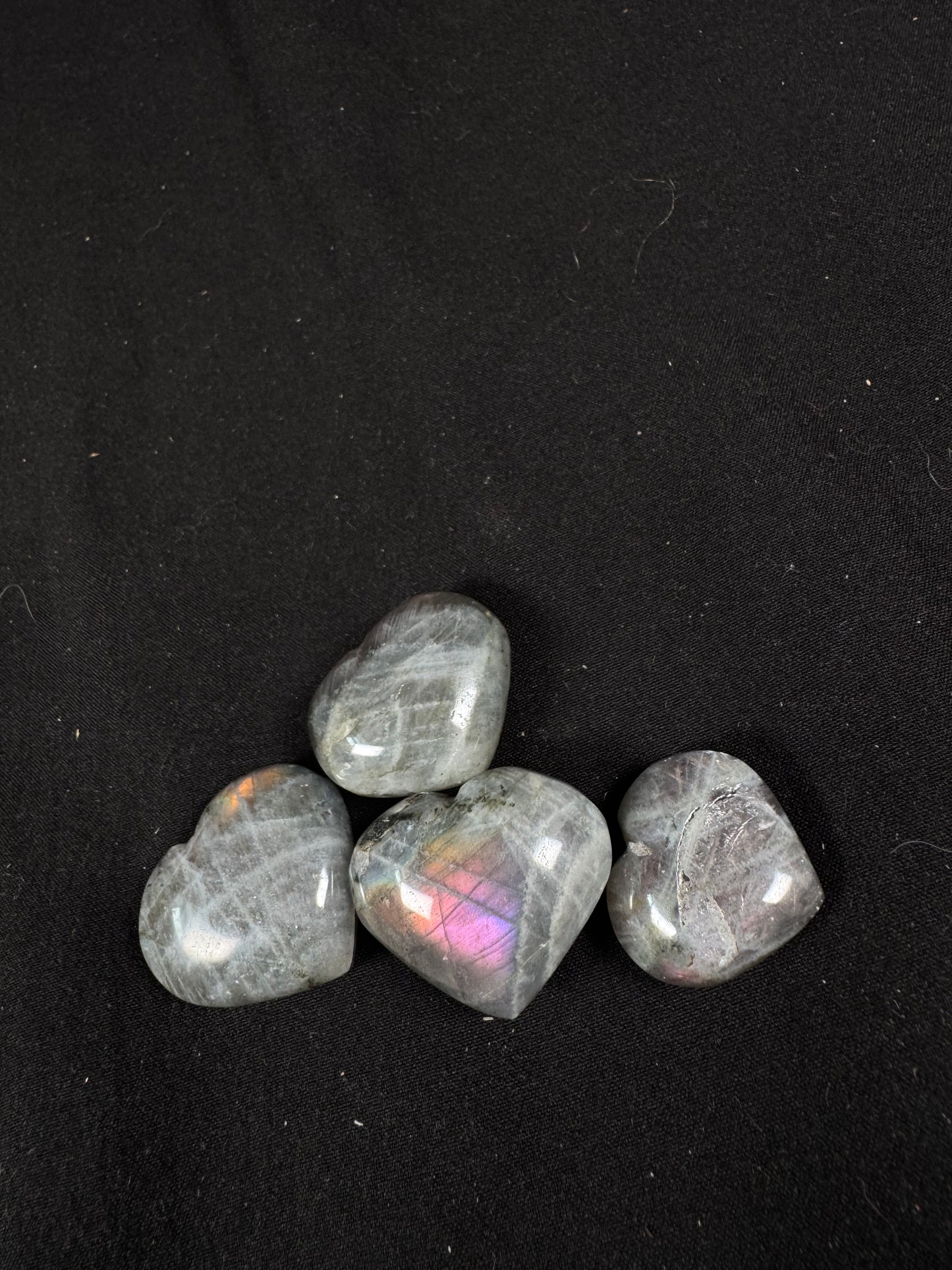 Polished labradorite heart carvings displayed on a black fabric background. Four gray hearts with natural black inclusions show flashes of colorful labradorite scenes, including vibrant pink, purple, orange, and subtle blue iridescence across their smooth, polished surfaces.