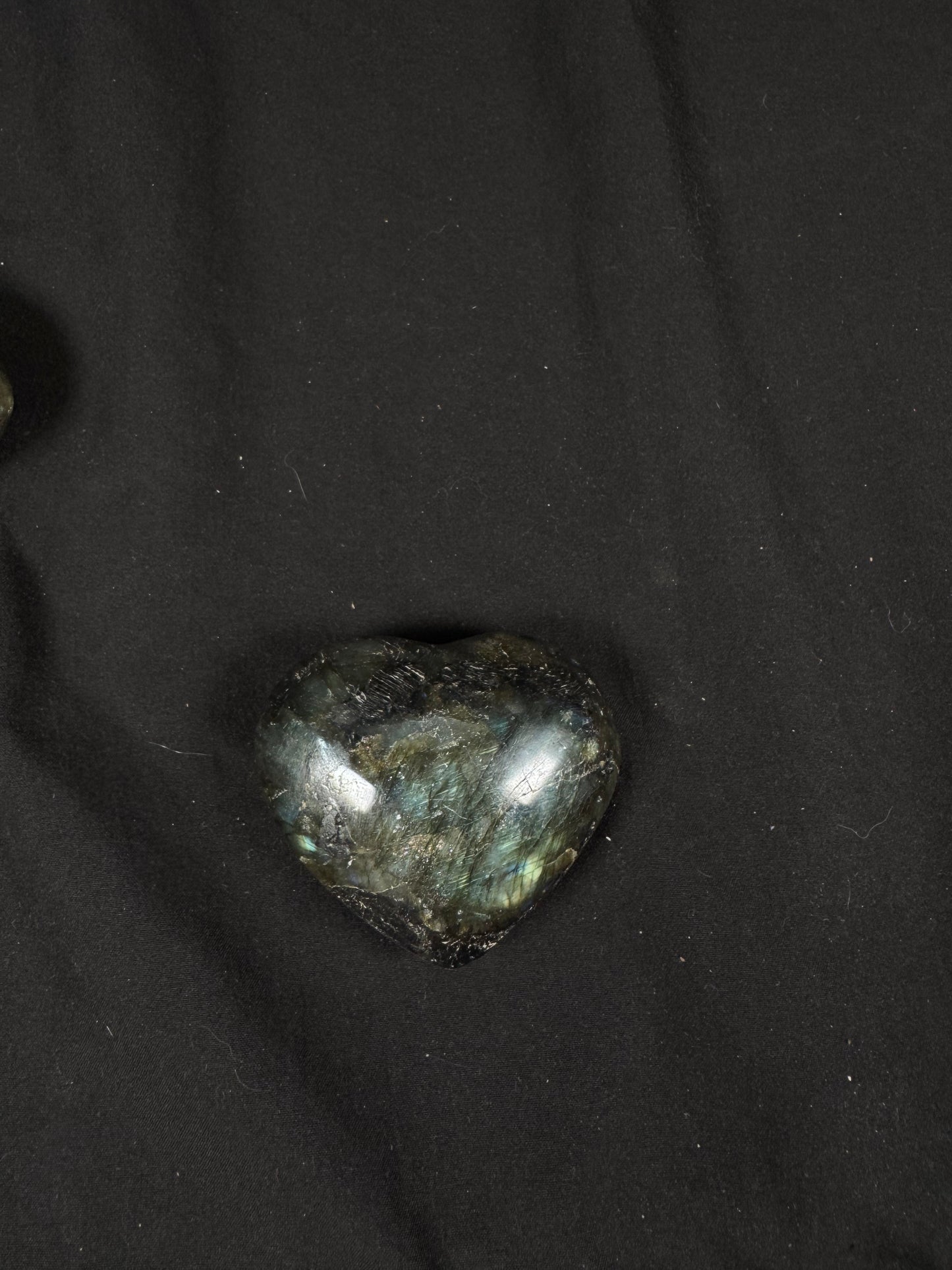 A single polished labradorite heart resting on a black fabric background. The dark gray to black stone features striking blue-green  with hints of gold, highlighting the natural patterns and glossy, heart-shaped finish.