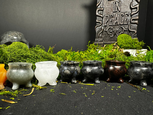  row of polished mini crystal cauldrons carved from a variety of natural stones, including milky white, gray, black, reddish-brown, and speckled crystals. The tiny cauldrons are displayed on a black cloth with vibrant green moss, creating a witchy apothecary aesthetic. A decorative skull and “Rest in Peace” tombstone prop appear softly in the background.