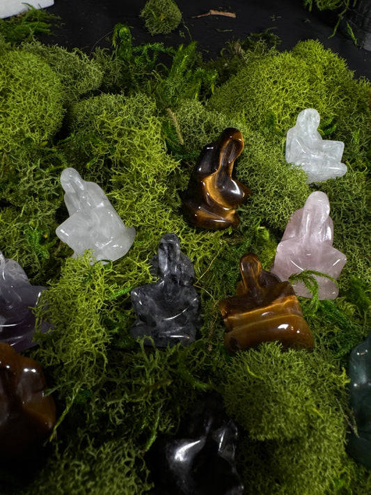 A collection of polished crystal Grim Reaper carvings displayed among vibrant green preserved moss. The assortment includes clear quartz, rose quartz, tiger’s eye, black labradorite, and other mixed gemstones, each carved into a seated hooded reaper figure with smooth, glossy details. The earthy moss backdrop highlights the variety of colors and natural patterns in each crystal carving.
