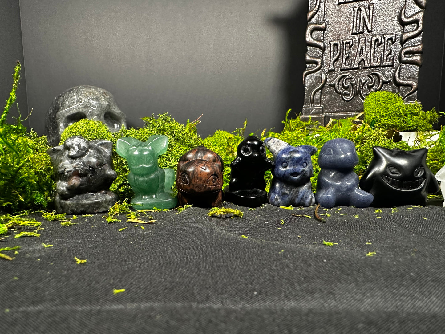 Seven miniature Pokémon-inspired crystal carvings arranged on moss against a gothic display. Featured characters include Snorlax, Eevee, Charmander, Gastly, Pikachu, Bulbasaur, and Gengar, each carved from a different natural gemstone with unique colors and patterns.