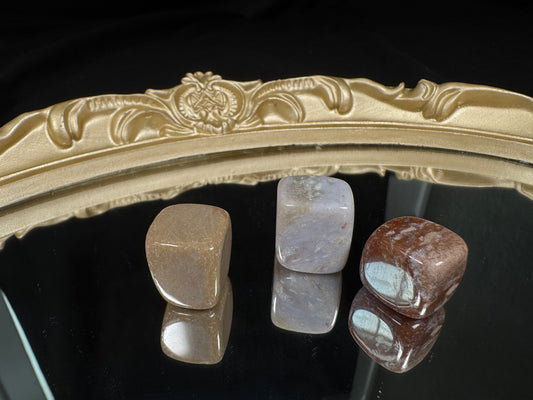 Polished Ocean Jasper cube tumble displayed on a black reflective surface with a decorative gold mirror tray. The cube features soft cream, beige, and muted blue-gray tones with natural orb-like patterns and subtle earthy inclusions. Its smooth, rounded edges and glossy finish highlight the unique colors and markings, while the reflective surface creates a mirrored view of the stone below.