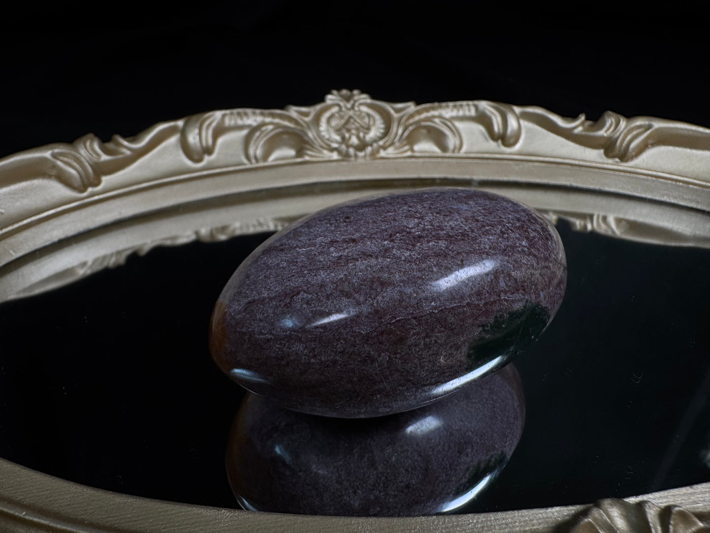 Polished Ocean Jasper palm stone with soft lavender and muted purple tones, featuring subtle natural marbling and a glossy finish, displayed on an ornate vintage gold-framed mirror against a black background.