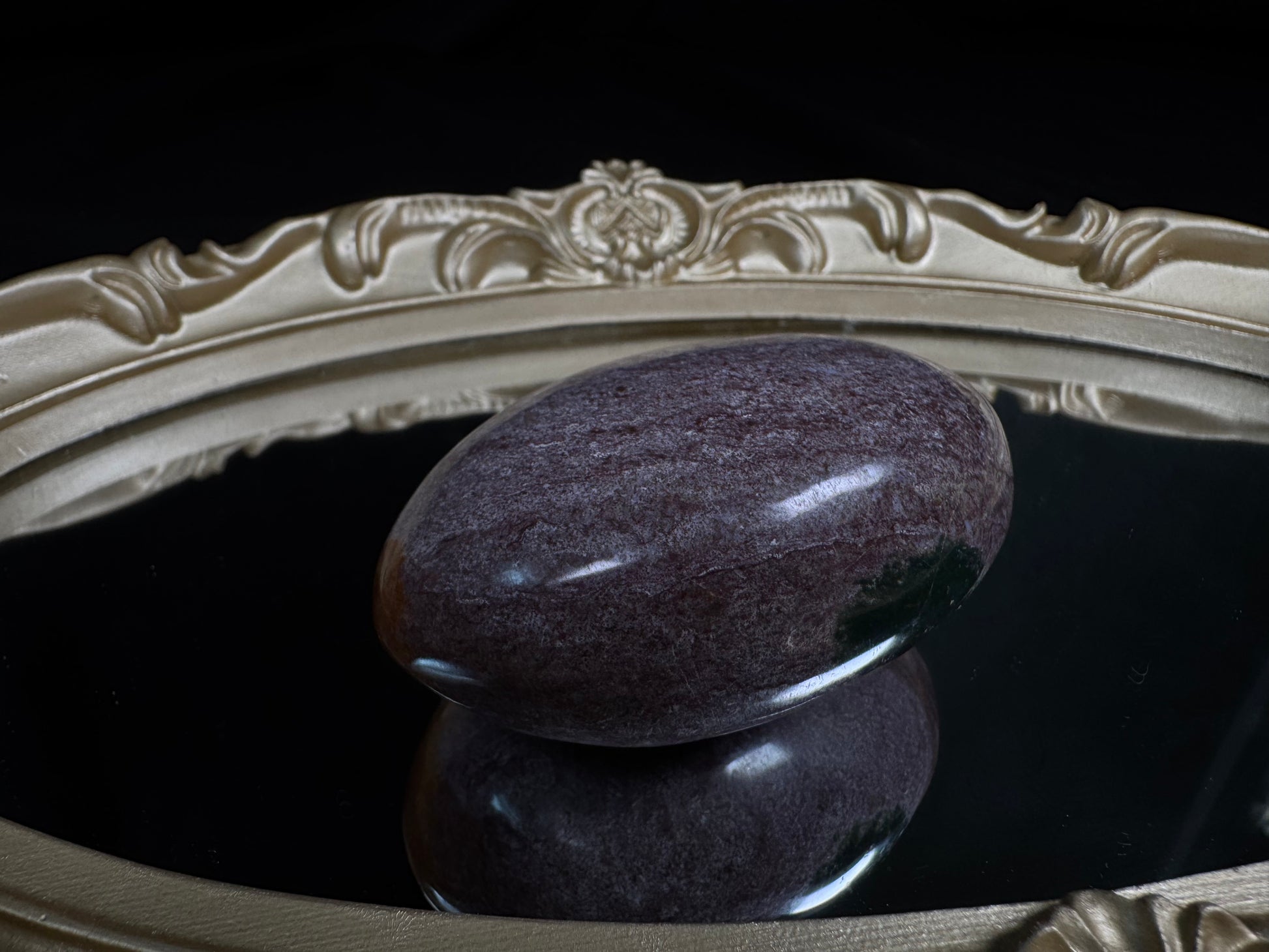 Polished Ocean Jasper palm stone with soft lavender and muted purple tones, featuring subtle natural marbling and a glossy finish, displayed on an ornate vintage gold-framed mirror against a black background.