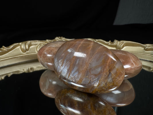 Polished peach moonstone palm stone with soft peach, cream, and translucent gray tones, featuring natural golden-brown veining and internal inclusions. The smooth oval stone is displayed on a reflective black surface with two matching peach moonstone palm stones resting behind it on an ornate vintage-style gold tray, creating an elegant, gothic-inspired presentation.