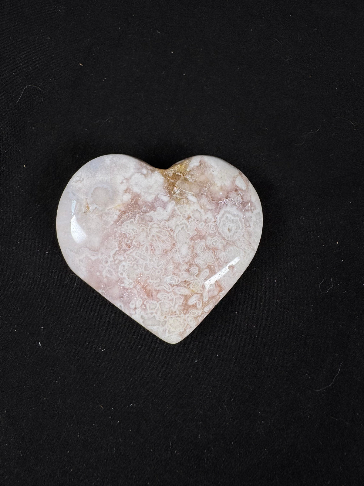 Polished pink amethyst heart displaying intricate lace-like banding in soft pink, white, and cream with hints of golden matrix near the top. The glossy heart showcases delicate natural patterns and subtle druzy texture, photographed against a black background.