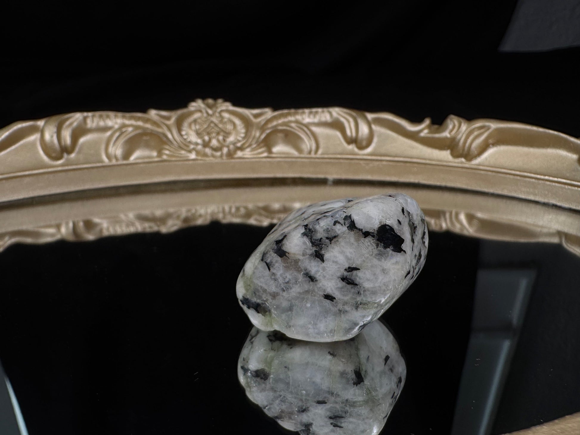 Polished rainbow moonstone tumble with black tourmaline inclusions standing on an ornate gold-framed mirror. The white stone features scattered black mineral inclusions and a subtle blue flash, reflected against a black backdrop.