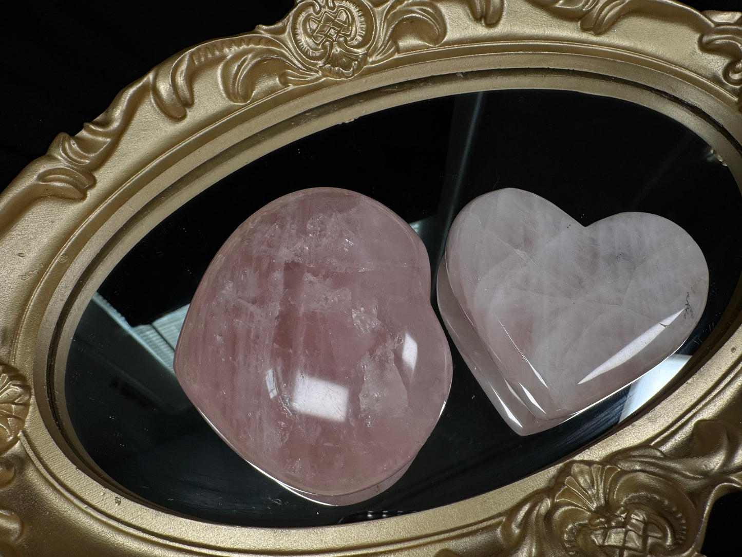 Two polished rose quartz hearts displayed side by side on a decorative gold-framed mirror. The larger heart is a deeper pink with visible natural inclusions and cloudy patterns, while the smaller heart is a lighter, milky pink with soft white veining and a glossy finish.