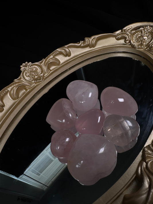 A collection of polished rose quartz hearts in soft shades of pale to medium pink displayed on an ornate gold-framed mirror. Each heart has a glossy finish with natural internal inclusions and subtle color variations, set against a black fabric backdrop.