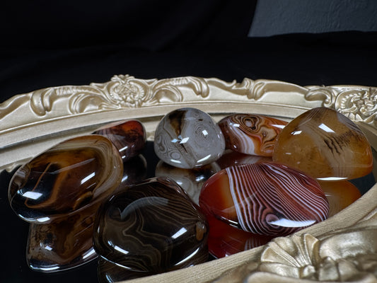 A collection of polished sardonyx tumbles displayed on an ornate antique-style gold mirror. The stones feature rich bands of deep brown, black, caramel, cream, gray, and reddish-orange, with striking natural stripes, swirls, and eye-like patterns. Their high-gloss finish reflects the light, highlighting the unique banding and smooth rounded shapes of each individual tumble against a black fabric backdrop.